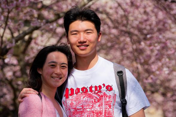 on the left an aisan woman with long black hair and a pink outer layer and to the right a taller asian man with a white shirt with red lettering and graphics and backpack straps has an arm around her shoulder -- pink blossoms in background