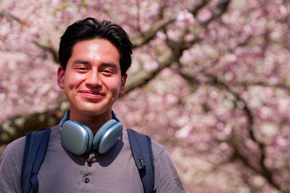 Man with moderate length hair and backpack straps and I think Airpods Max smiles in front of pink tree blossoms
