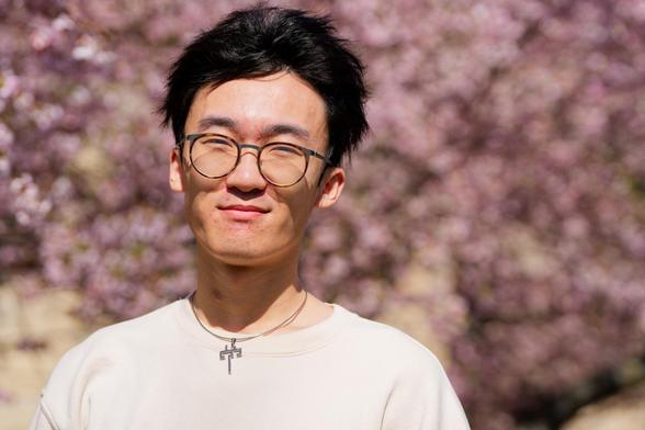 Asian man with white shirt and round glasses has a "^ ^" expression, white shirt and a small necklace in front of blurred out pink blossoms