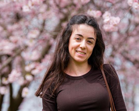 Woman with hair past the shoulder and brown blouse with pink tree flowers against a bluish background