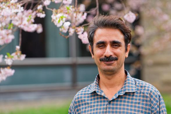 South Asian or Middle Eastern man with mustache and neat short hair with a checkered blue and yellow button down shirt with a few blurry pink tree flowers behind and a big expanse of window behind
