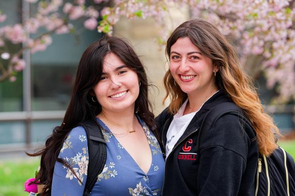 Dair haired women on the left with blue blouse with flowers and lighter haired owman on the right with a dark jacket with a CORNELL logo.  Both wearing backpacks.  Flowers near the top adre in the background and a glass, steel and stone building behind them.