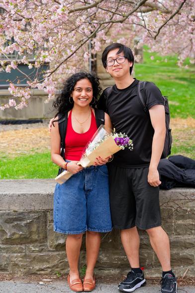 Woman on left with red blouse, boquet of flowers and curly hair and denim knee-length shorts and a man on the right with glasses, short dark hair, dark shirt and dark pants with the same length as the woman.  behind them is a short stone wall,  tree flowers and a bit of building.