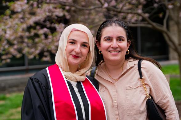 Women on the left with headscarf and red painted lips and black robe with crimson strips and on the right with braided hair and beige shirt with green grass flowers and windows behind