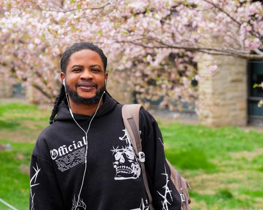 Black man with black shirt, earbuds and backpack in front of treelawn