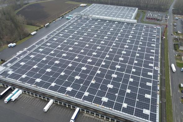 An aerial view of a large industrial building with a roof covered in solar panels. Trucks are parked alongside the building and on the adjacent road, surrounded by grassy fields and trees in the background.