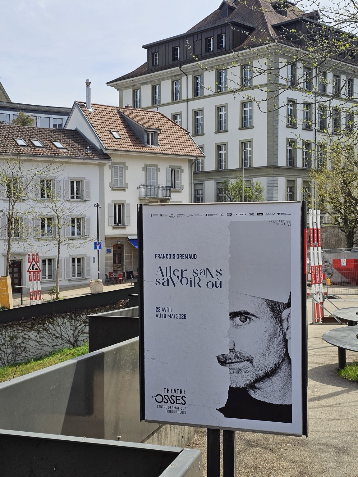 Une affiche du spectacle « Aller sans savoir où » en ville de Fribourg. On aperçoit le lycée du Collège St-Michel, et on se rend compte que l’affiche est posée devant la Bibliothèque universitaire. Sur l’affiche le visage de François est coupé en deux.  . 