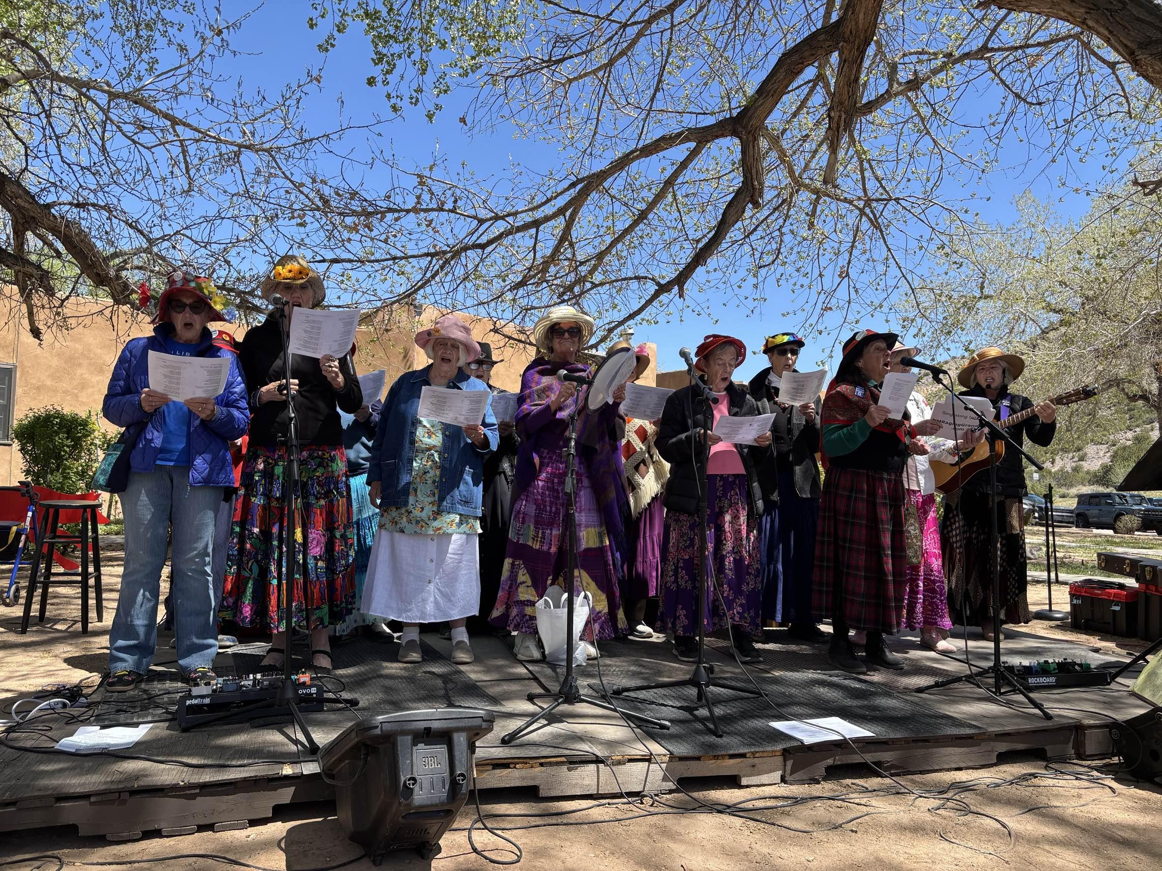 Group of women singing on a stage outdoors. They are in an array of hats and colorful clothes.