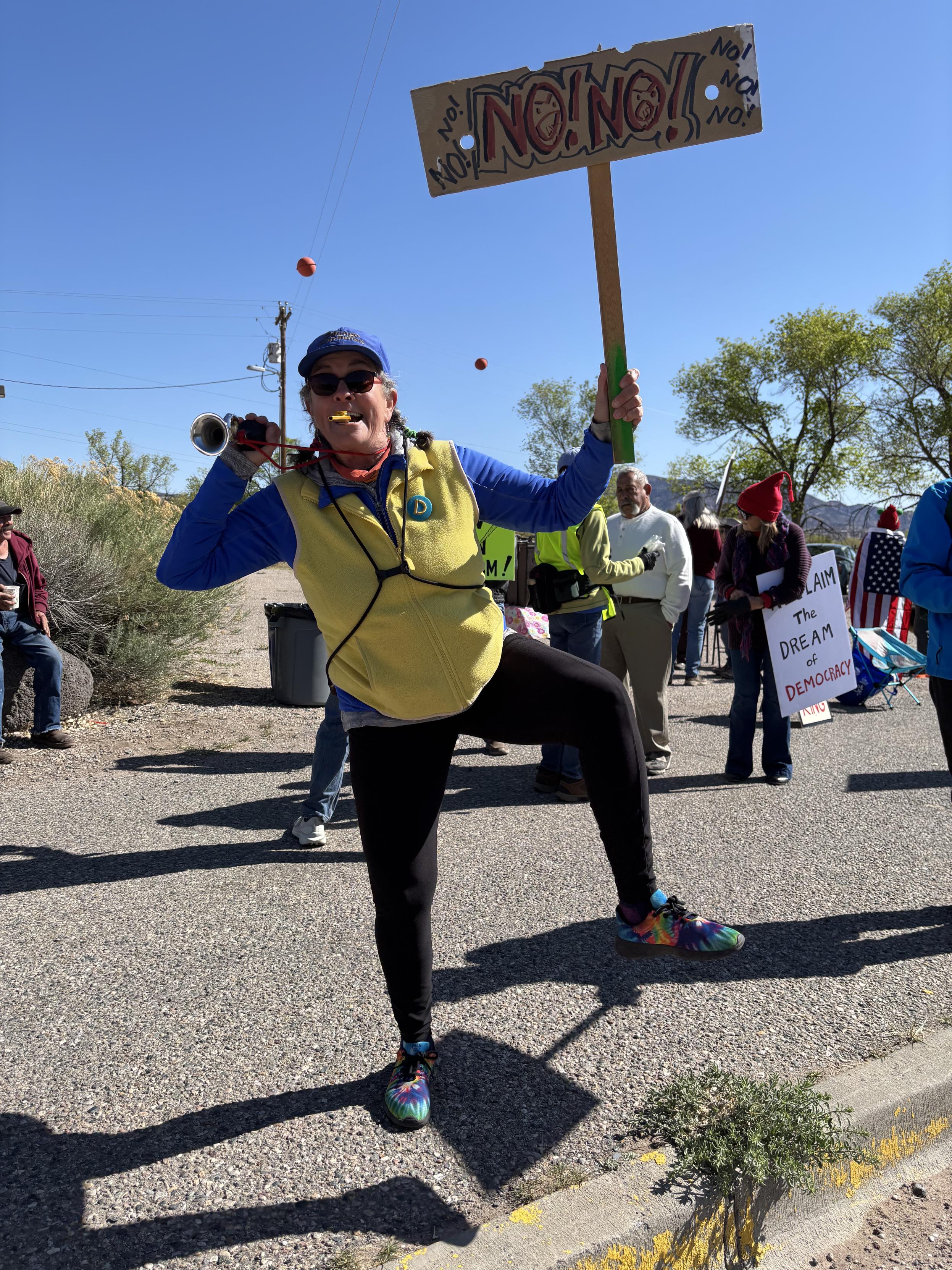 Woman holding a sign reading No! No!, holding a horn, playing a kazoo, wearing a bright yellow vest and multi-colored shoes.