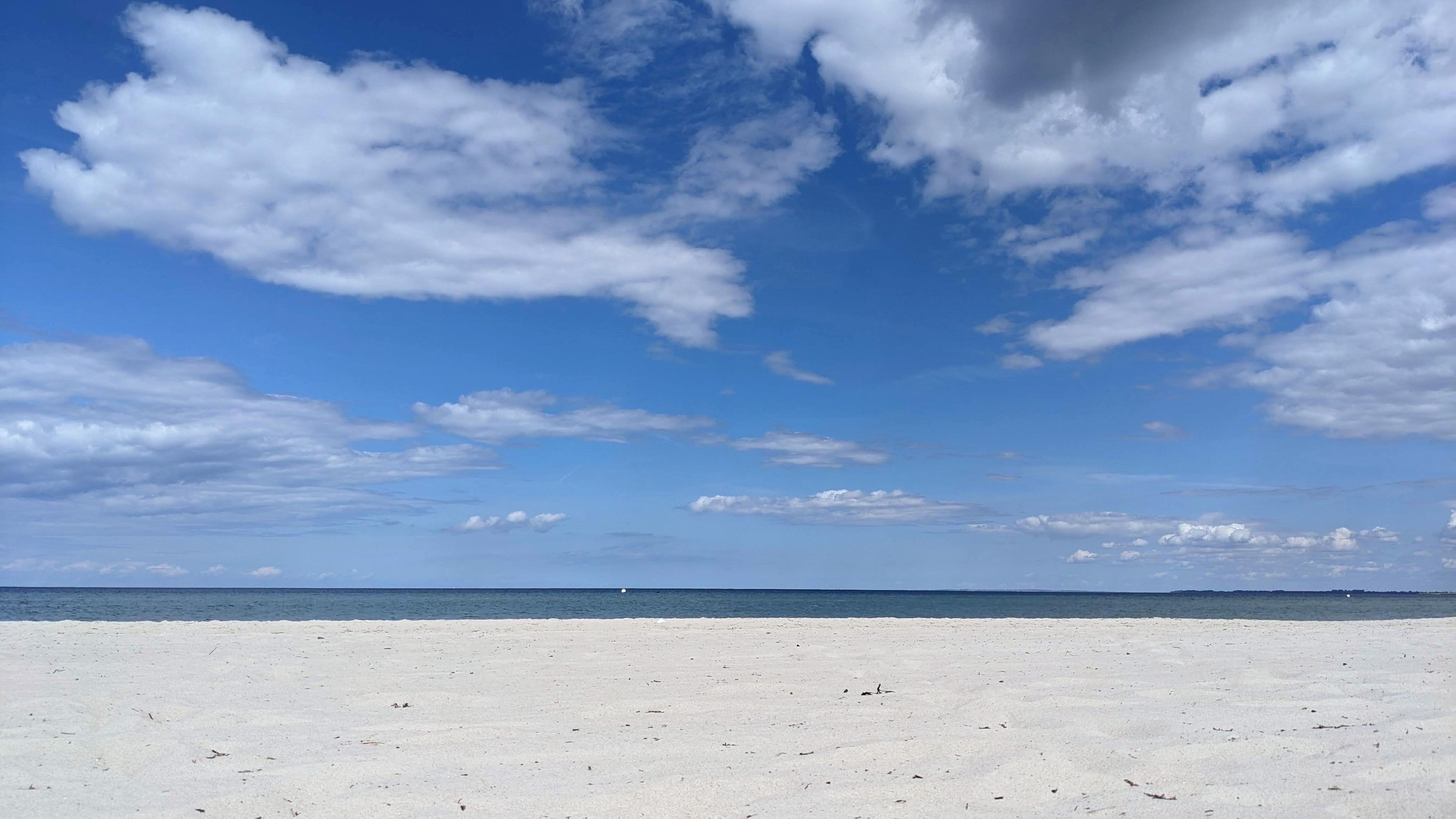 Bildbeschreibung von KI:
Ein weiter leerer Sandstrand erstreckt sich im Vordergrund. Der Sand ist hell und fein und wirkt glatt mit nur wenigen kleinen Spuren. Dahinter liegt das ruhige Meer der Ostsee. Das Wasser ist dunkelblau und bildet eine gerade Linie am Horizont.

Der Himmel nimmt einen großen Teil des Bildes ein. Er ist leuchtend blau und von vielen weißen Wolken durchzogen. Einige Wolken sind groß und weich geformt andere kleiner und verstreut. Das Licht wirkt klar und freundlich wie an einem ruhigen Tag ohne Sturm.

Am rechten Rand in der Ferne ist eine flache dunkle Landlinie zu erkennen die leicht in das Meer hineinragt. Auf dem Wasser ist weit draußen ein kleines helles Objekt zu sehen das wie ein entferntes Boot oder eine Boje wirkt.

Das Bild vermittelt eine ruhige offene Atmosphäre mit viel Weite und wenig Bewegung. Es zeigt die stille Landschaft der Ostsee mit Strand Meer und Himmel in klaren einfachen Formen.
