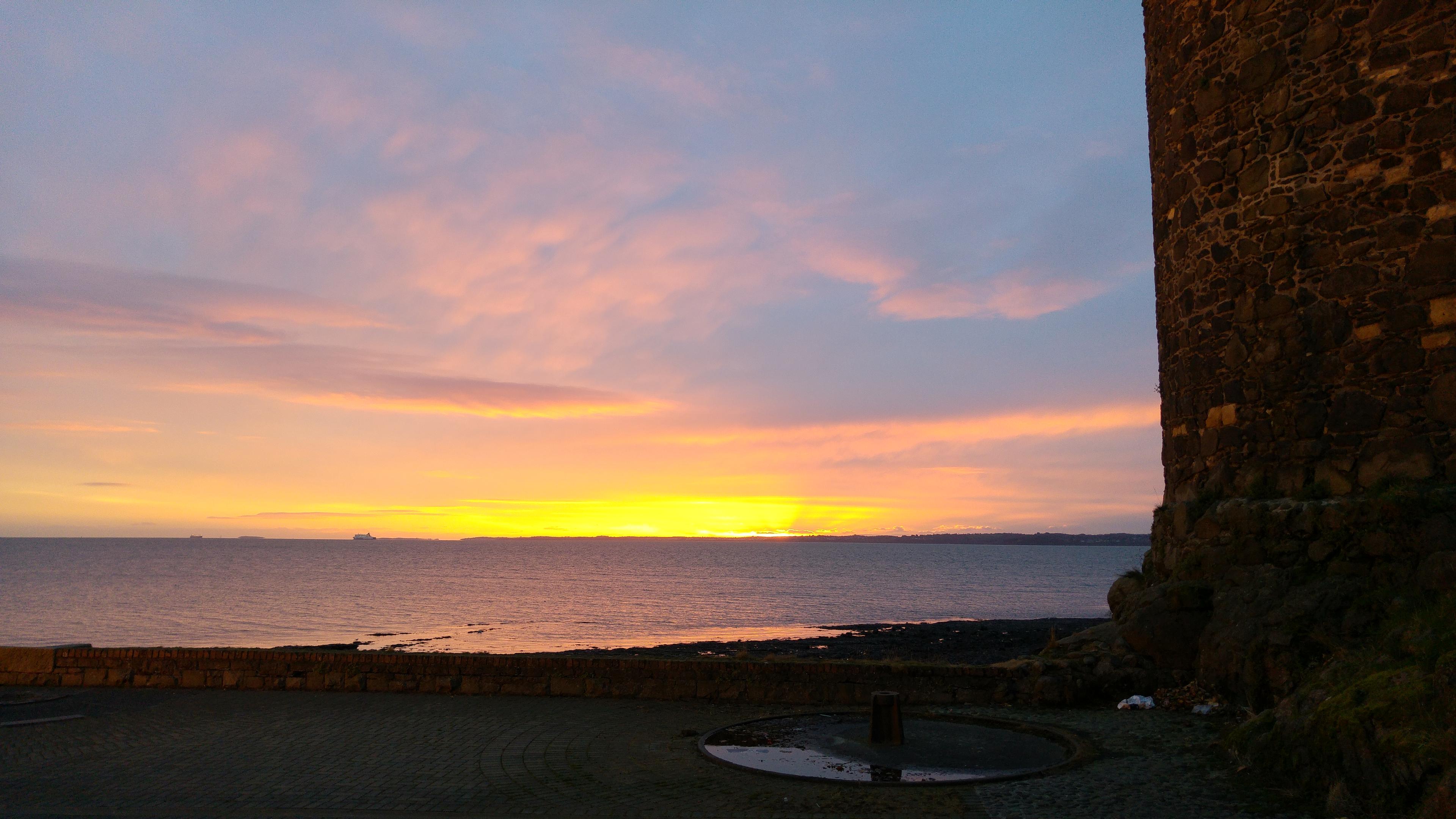 Sun rises over Belfast Lough, the sun breaking through some light clouds. The seascape is framed on the right by the (slightly askew) edge of Carrickfergus Castle, and in the foreground are the shallows of a small beach. In the distance, on the horizon, is a ferry.