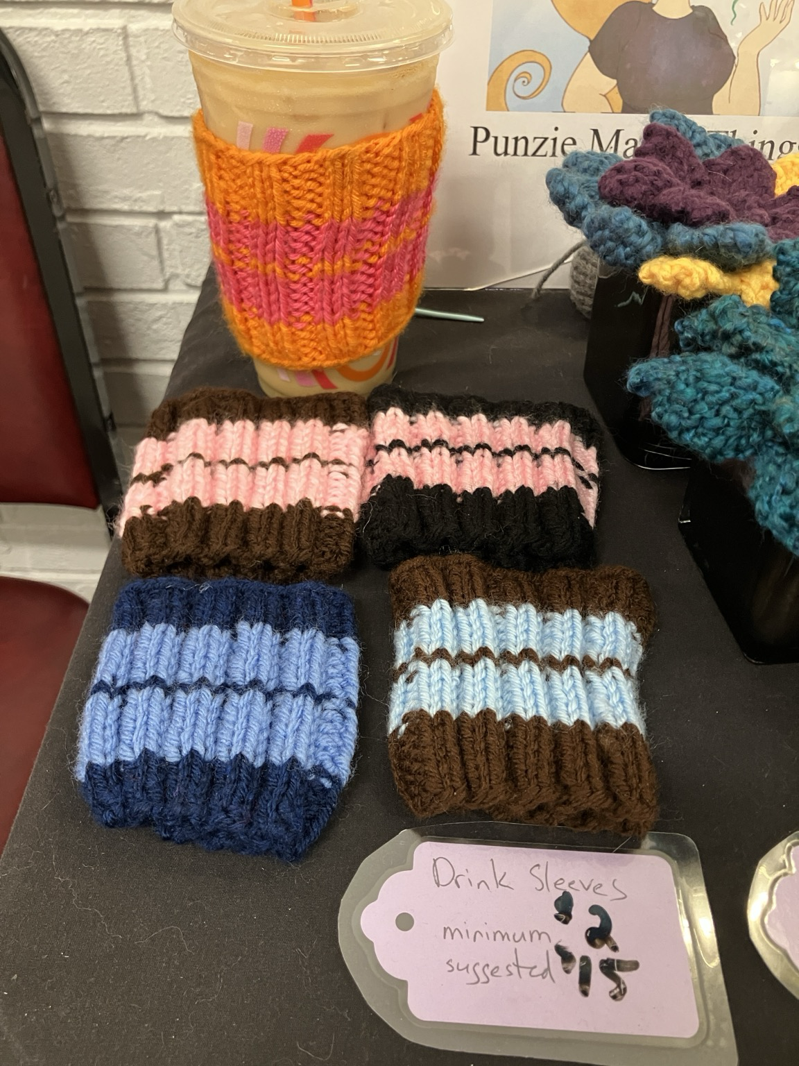 A Dunks iced coffee in a pink and orange knit drink sleeve. On the table in front of it are knit drink sleeves in different colorways