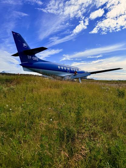 A small blue and white airplane is parked in a grassy field at Reykjavík’s airport under a partly cloudy sky. TF-ORD is a BAe Jetstream 32 registered to Eagle Air Iceland. 