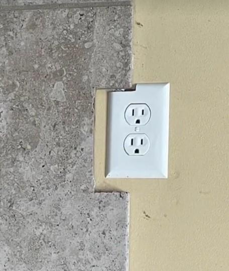 An upside down picture of plug sockets badly fitted to a wall. A tile has been cut to accommodate the sockets but it doesn't line up, so the corner of the sockets has been cut off to accommodate the tile. The three holes in upside down sockets look like faces with worried expressions.
