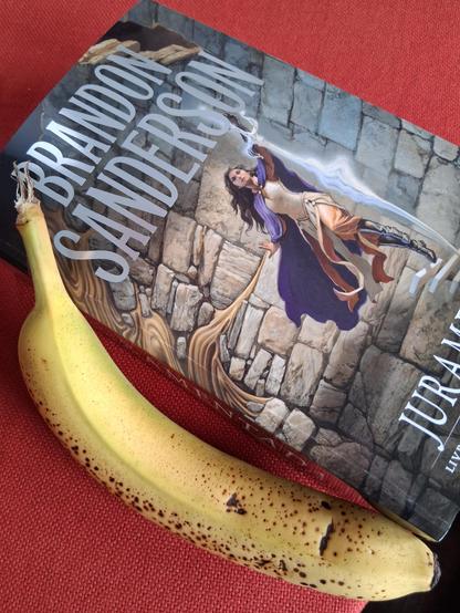A large book reading "BRANDON SANDERSON" and "JURAMENTADA" in its cover is over a red cloth and next to a banana for scale. Here, it's viewed from the top.