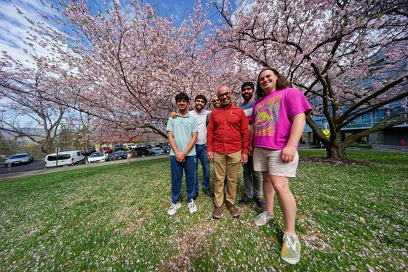 Five men, mostly South Asian, stand together under two spreading cherry trees on a grass lawn with scattered petals.
