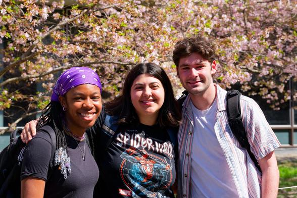 Three students left to right: a black girl with a purple bandanna, a lighter skinned woman with dark hair and a dark spider-man shirt, and a man with a white undershirt and striped open button down on top.  Sakura trees above look like they are a little bit past their peak