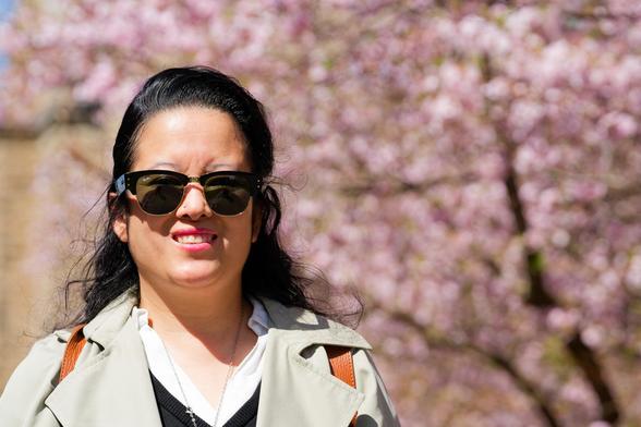 Woman with dark hair and dark glasses wearing a light colored outer layer and something black and white underneath with blurry pink flowers behind
