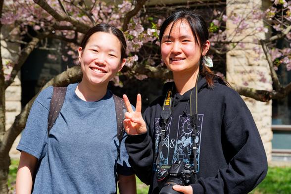 Left: an asian girl with a blue t-shirt and dotted backpack straps leaning towards the center; Right: another asian girt with a dark hoodie and a Nikon making a v-sign;  behind find cherry trees past their peak and the facade of Olin Library