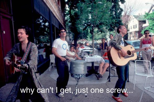 The Violent Femmes performing outside at Tuts with the subtitle "Why can't I get, just one screw..."