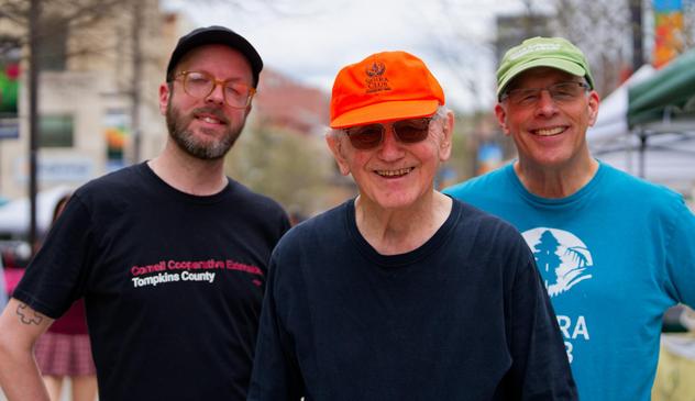 With the ithaca commons as a backdrop: three men with billed caps,  left to right: beard black shirt and cooperative extension logo, dark shirt and orange sierra club cap and green cap and blue Sierra Club Shirt