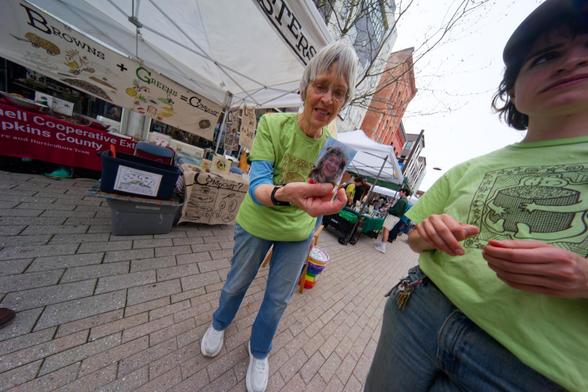 Grey haired woman wearing a green shirt holds a picture of a man (me!  wait a second,  who are you calling a "man"?) with a younger woman in a green shirt partially visible to the far right and a composting booth on the left