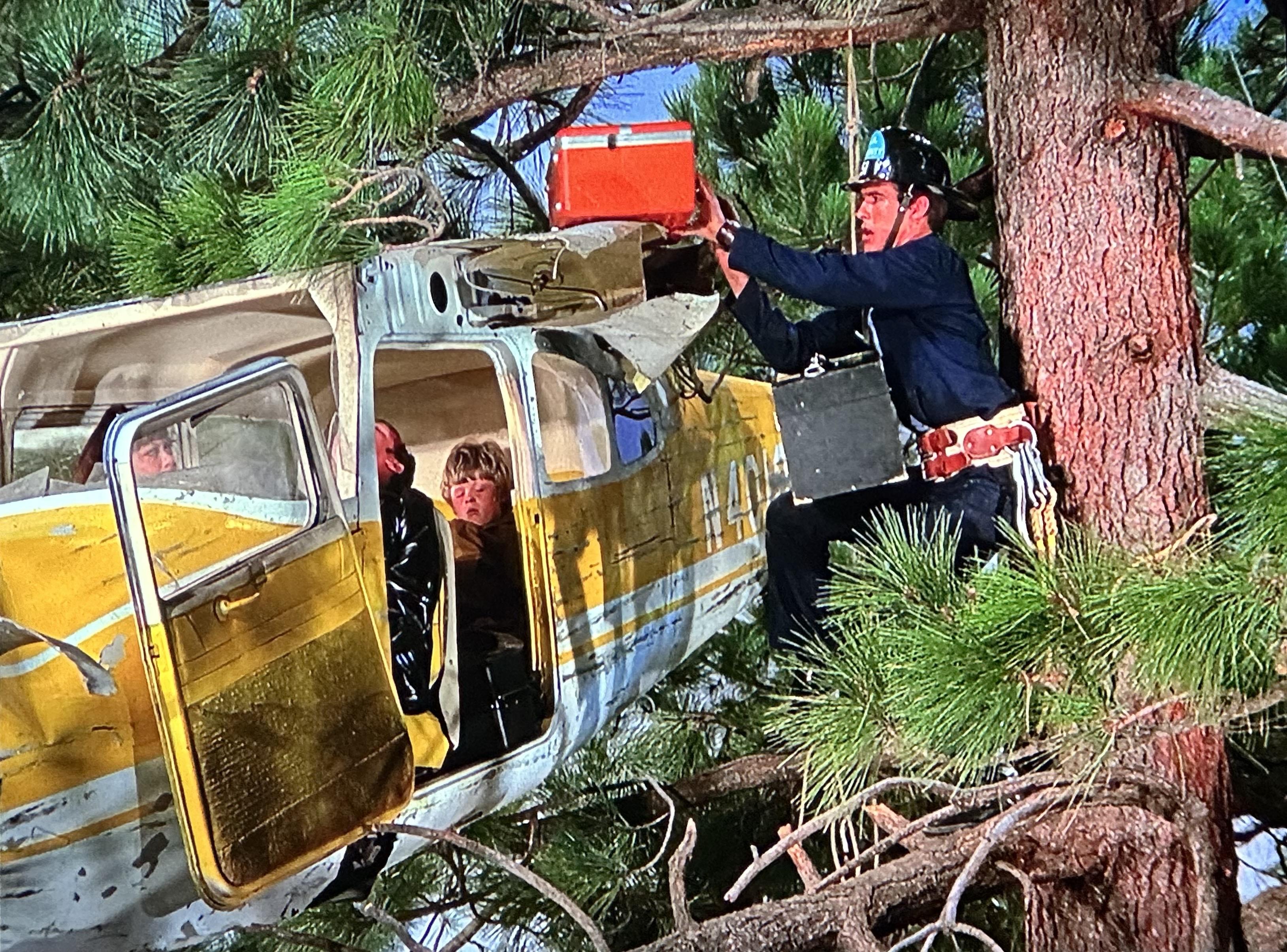 Fireman gauge from the TV show emergency precariously resting his equipment on top of a small crashed Sesna plane that is perched a top a 100 foot pine tree deep deep in the forest. An injured man woman, and child sit inside.