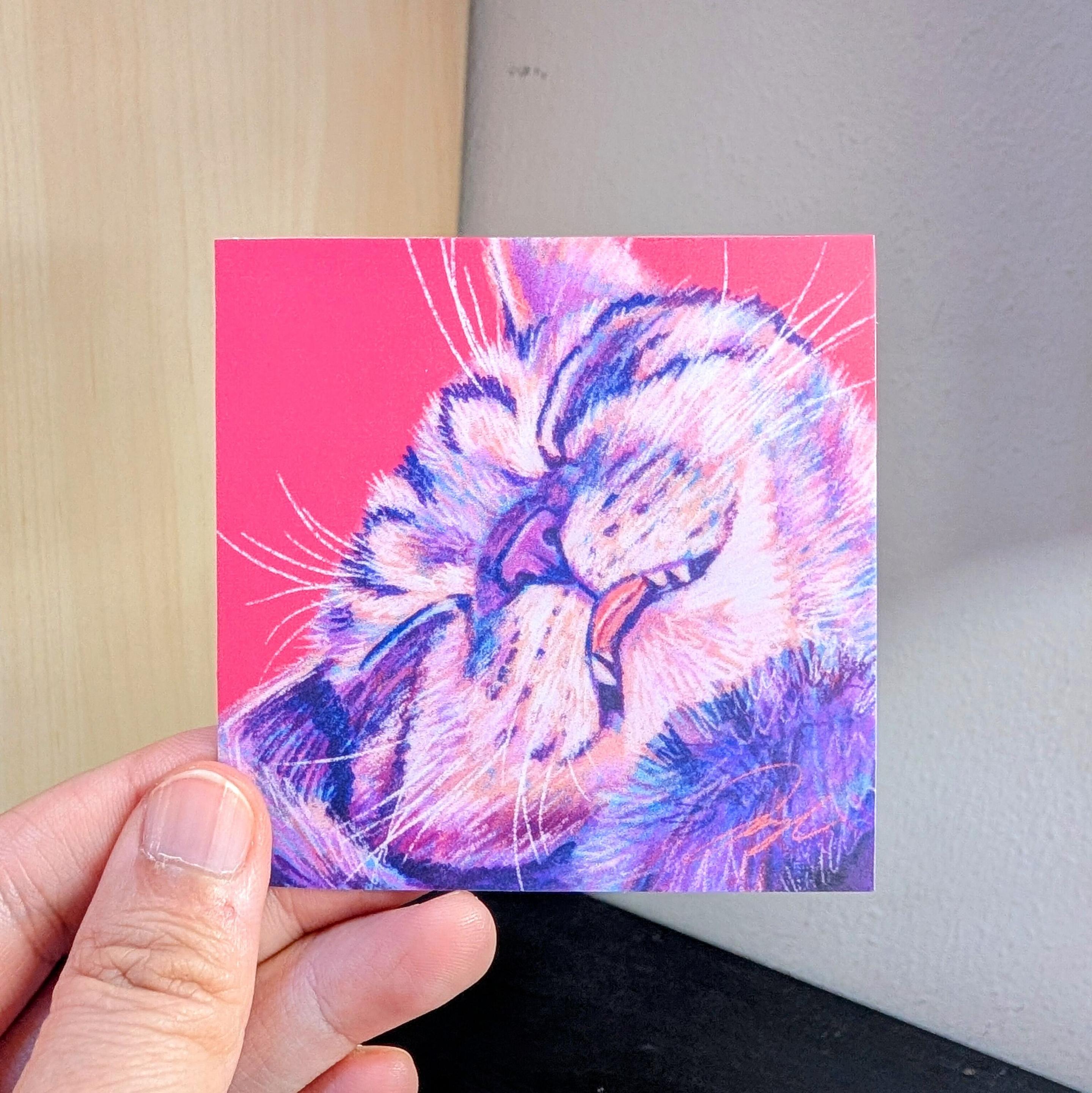 Photo of a left hand holding a square sticker. The sticker is a matte painting of a sleepy cat.