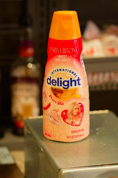 Paris Hilton branded coffee creamers with a dark tan top red and pink label: Paris Hilton, Sweet and Spicy, International Delight with a stylized sun between those words and half a coffee cop seen from the top, That's Hot, Coffee Creamer, 32 fl oz 1 qrt.  it rests on a rectangular box which is dirty from creamer droplets and something blurry in the background that looks like an alcoholic beverage but it isn't as well as other paraphrenalia of a poorly thought out condiment station
