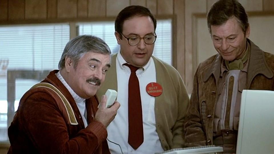 Scotty speaking into an old Macintosh computer mouse while Dr. McCoy and Dr. Nichols look on from Star Trek IV the Voyage Home