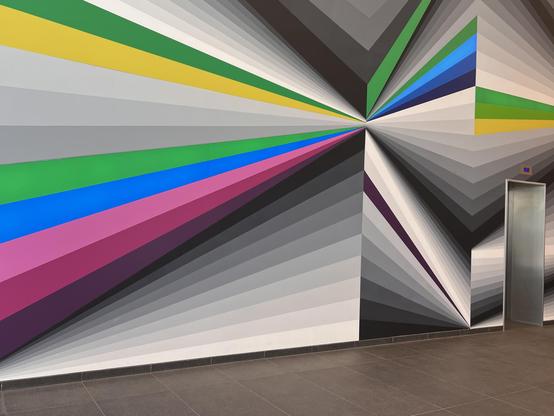 Op-Art-Wandgemälde in der Eingangshalle der Sparkassenakademie Stuttgart. Streifen in bunten Farben (u.a. Grün) und Grautönen laufen aufeinander zu, was einen dreidimensionalen Eindruck erweckt. Am rechten Bildrand eine Fahrstuhltür, unten angeschnitten der Fußboden.