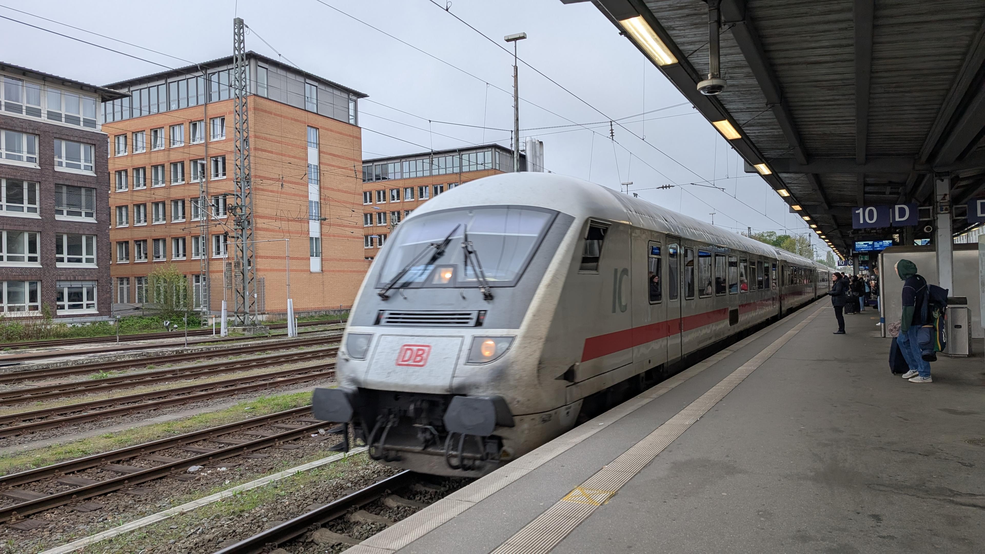 A DB IC 1 train with control car at the front of the train arriving at the platform, some waiting passengers on the platform, office buildings behind the train tracks