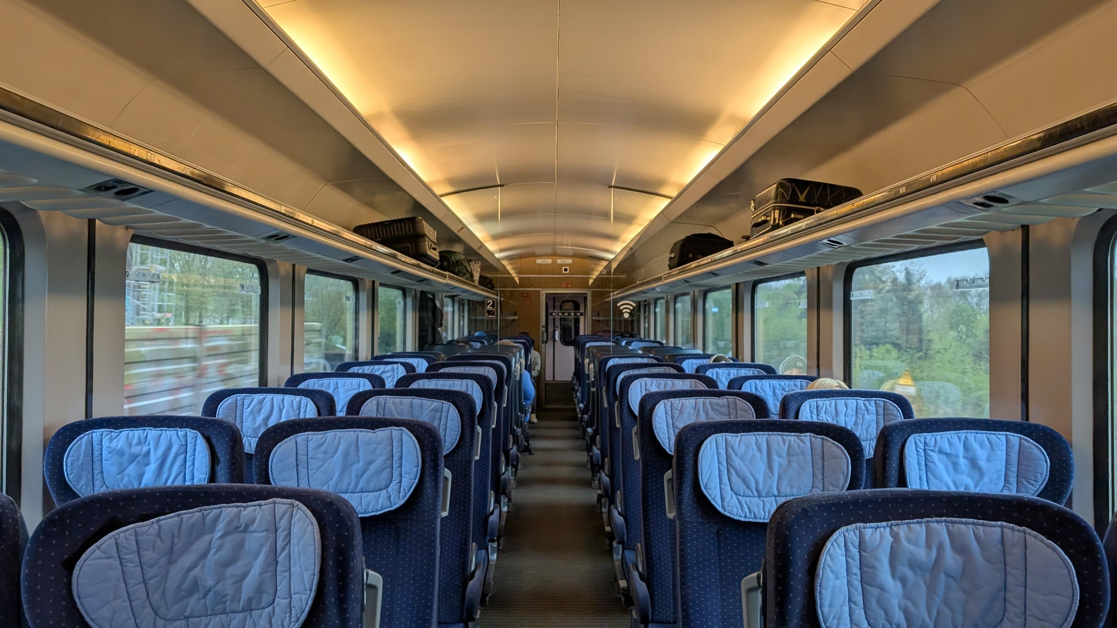 Interior of the IC 1 coach, 2+2 seating configuration, blue seats, light-blue headparts, wooden-like panelling at the end, only few passengers