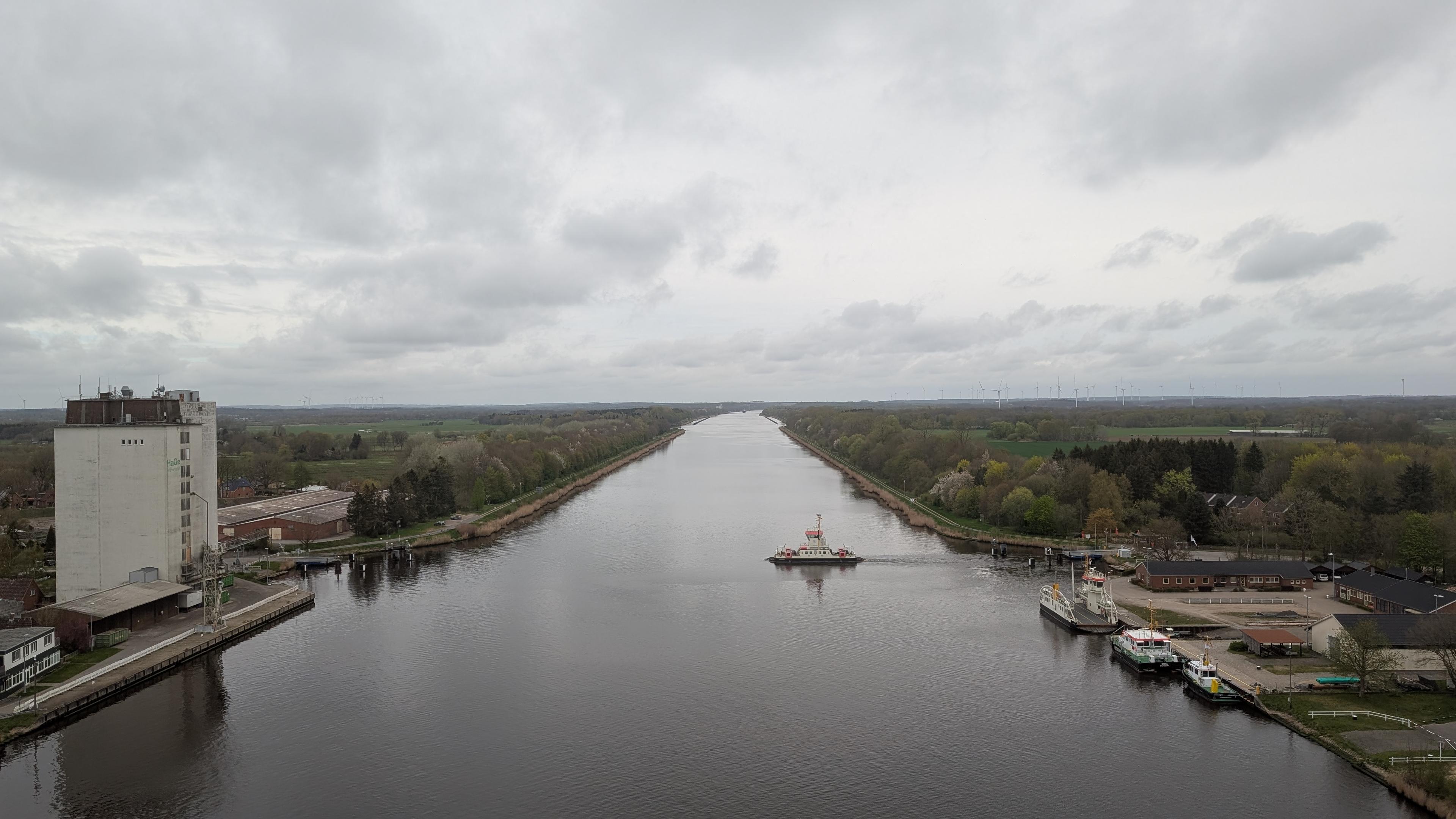 View of the Nord-Ostsee-Kanal from above, a ferry crossing the canal, a high industrial building at the left, many parked boats at the right, dark cloudy weather