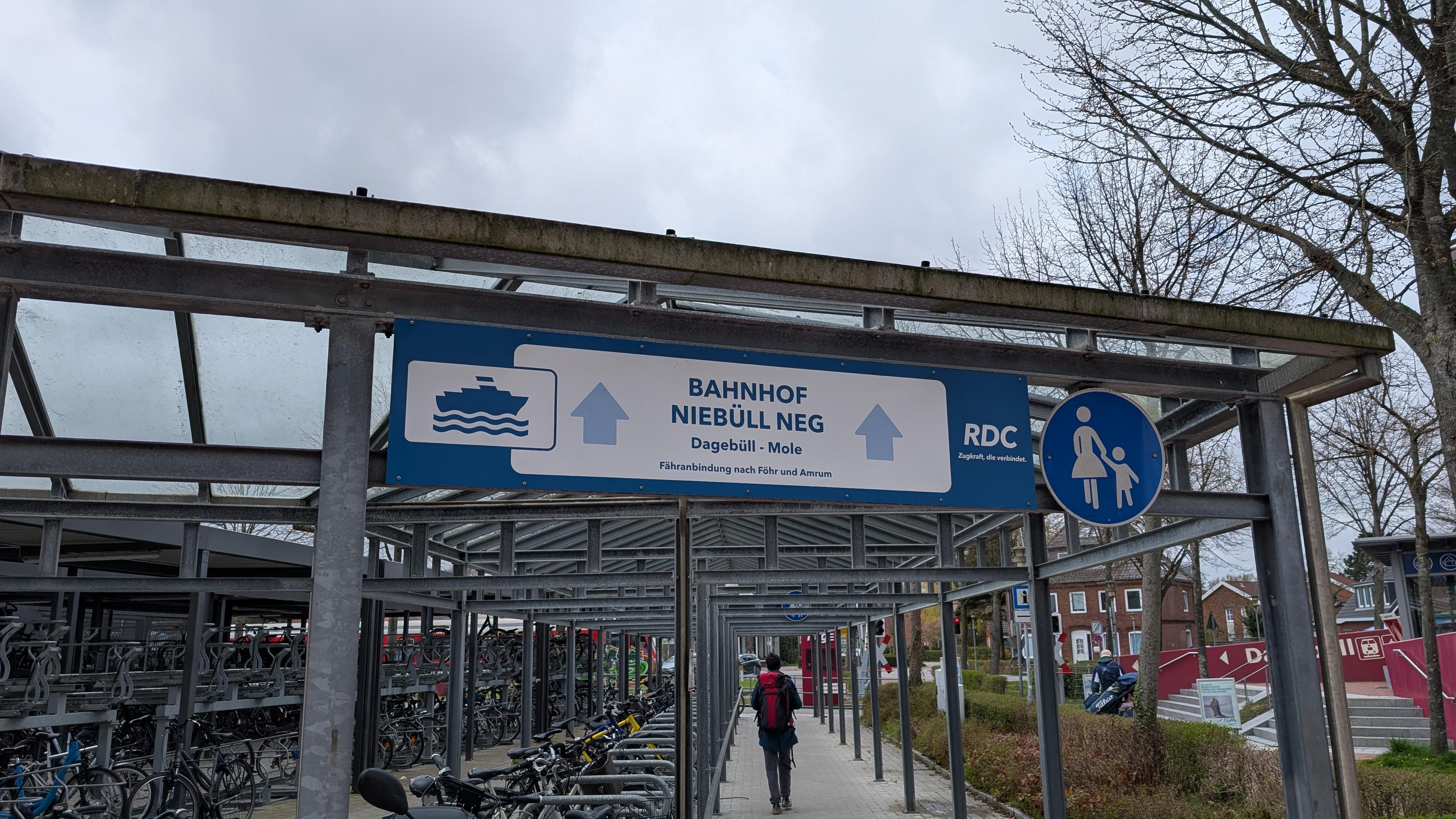 Roofed passage with double-deck bike stands, an overhead sign pointing towards "Bahnhof Niebüll neg" with a ferry symbol and an RDC logo