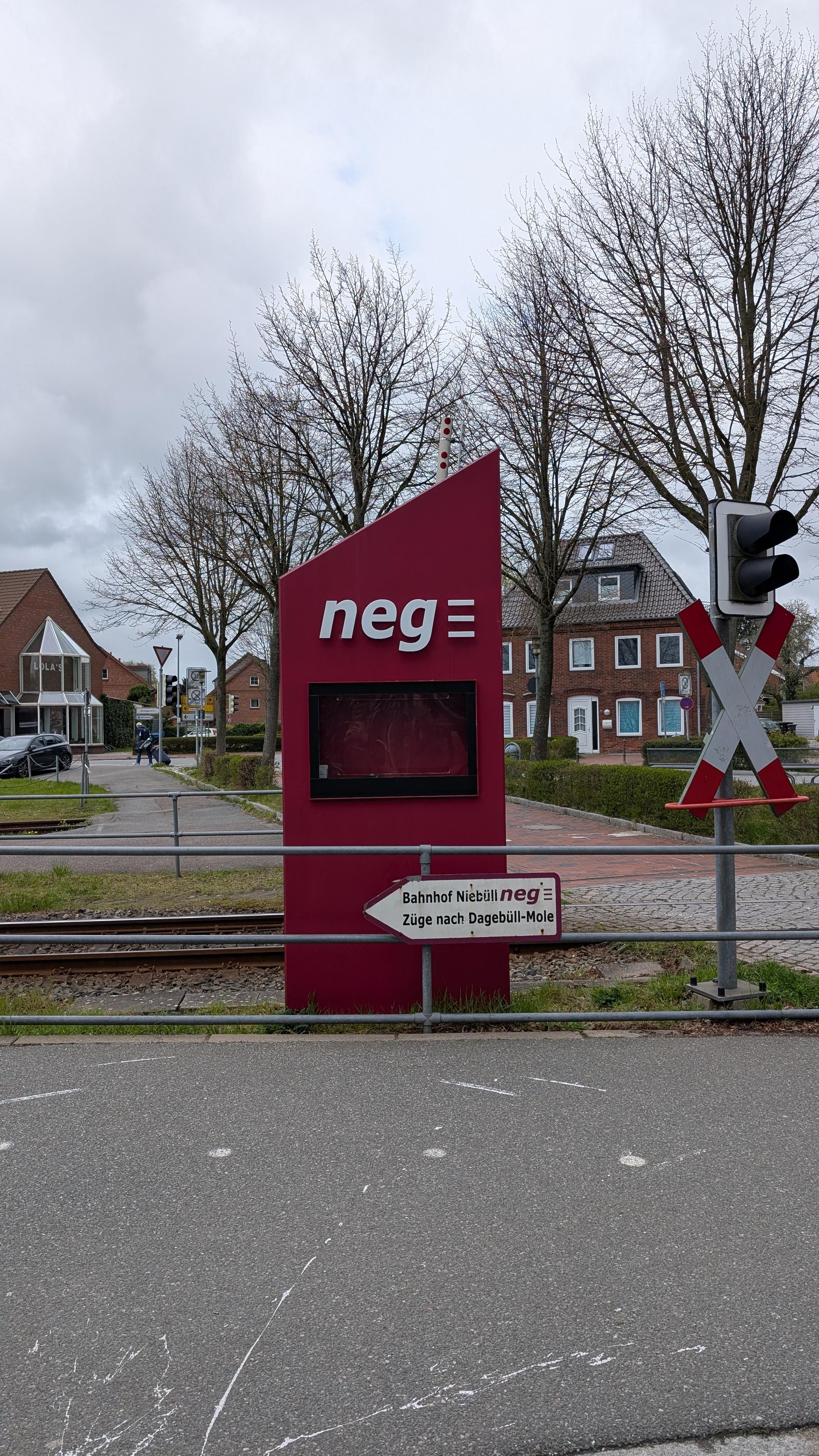 A red information pillar, empty display, big neg logo, a signpost with "Bahnhof Niebüll" and neg logo pointing to the left, "Züge nach Dagebüll Mole", a track and a level crossing in the background