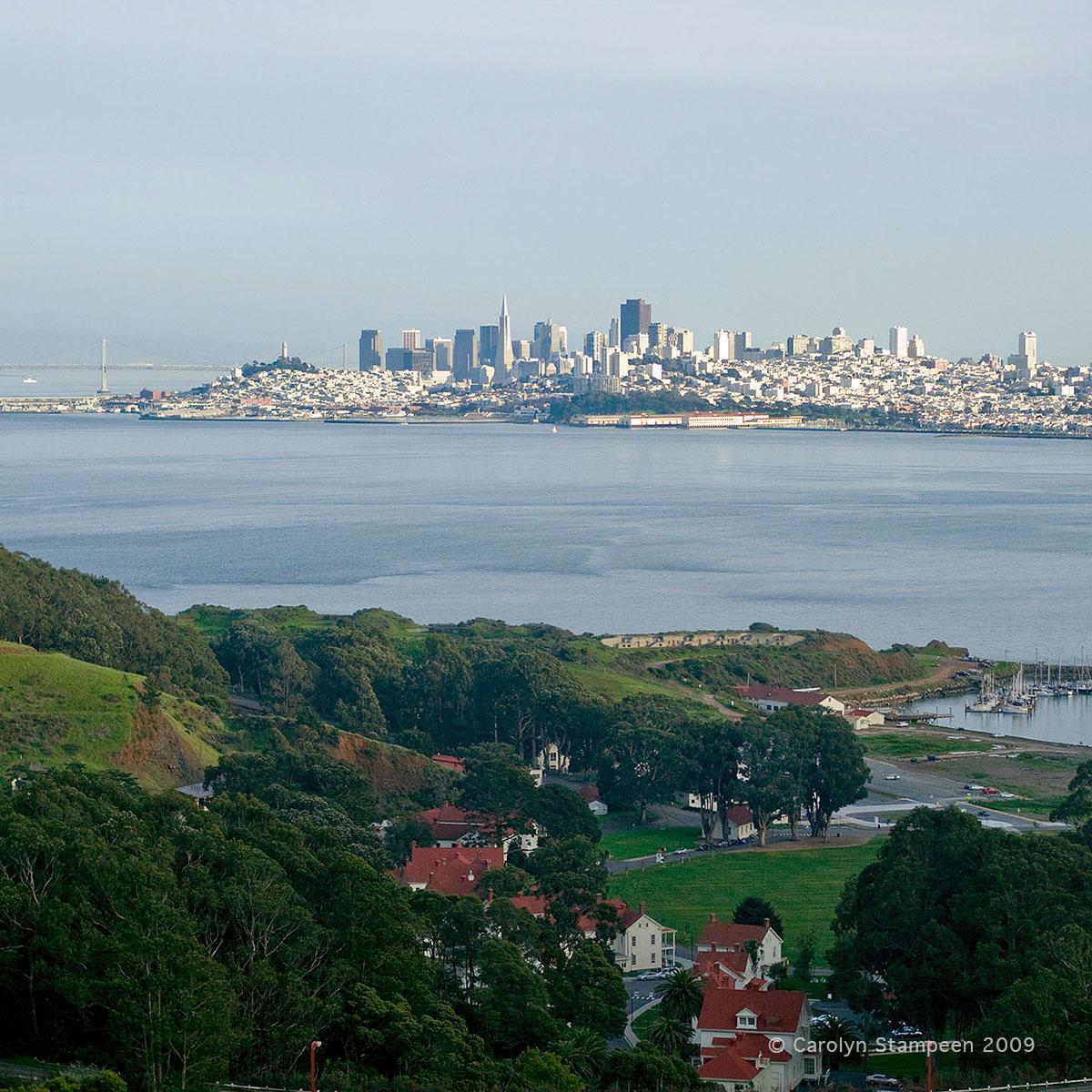 Taken from a bus, on the other side of the Golden Gate Bridge, before entering the tunnel.  We have a great view of San Francisco, with the Bay Bridge visible.  Below us, we see green trees and grass below us, as well as a marina and white buildings with red roofs.