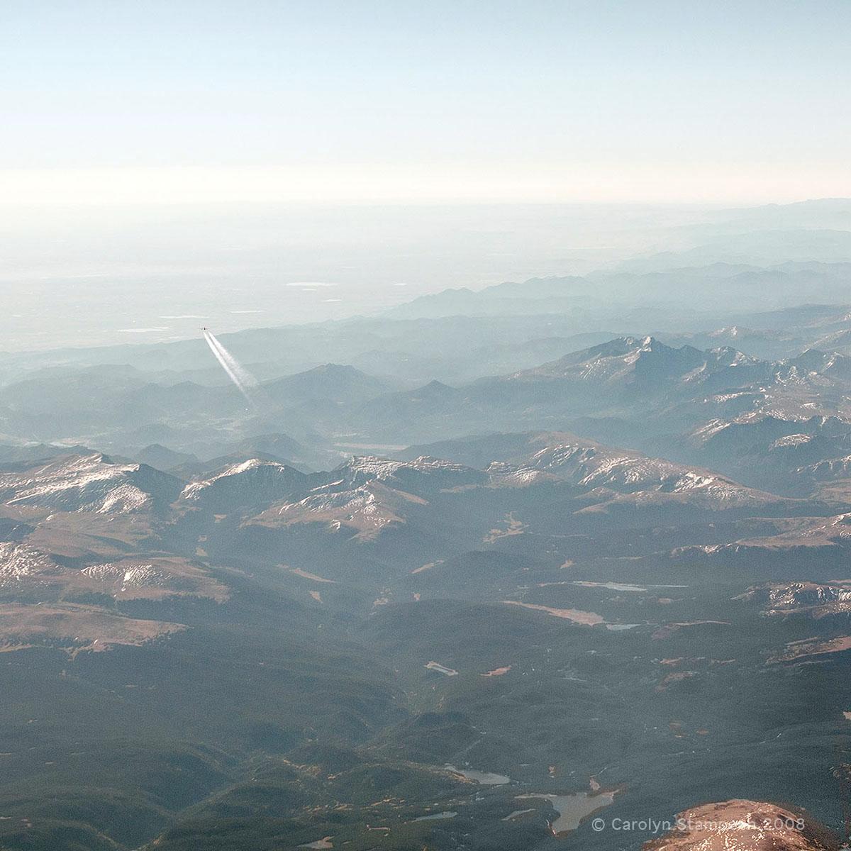 We are looking down on snow-capped mountains and green river valleys.  And...flying away from us is another plane, contrails streaming behind it, about to disappear in the haze.