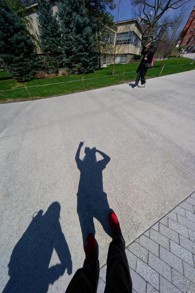 Stretched out shadow of person wearing fox ear headband making the kitsune hand gesture on a sidewalk with another person's shadow behind, a distant walker and a building with evergreen trees in front behind her