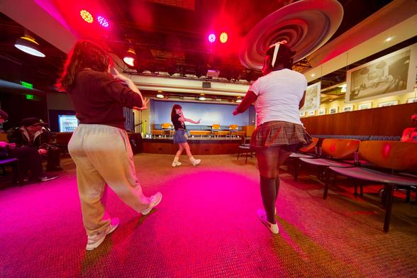 Close to mean find a white girl and a black girl looking away from me when dancing while purple and pink lines shown down on use and a further away white girl steps to the left with an outstretched hand