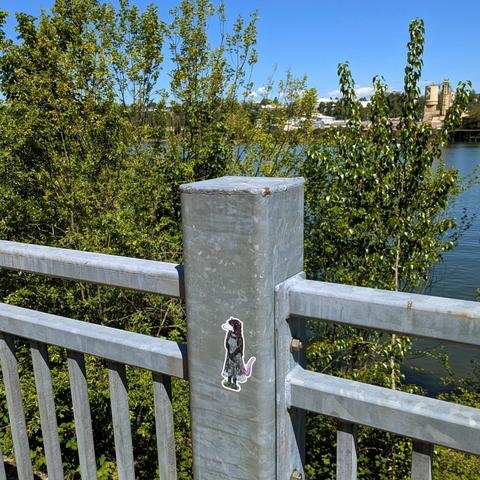 Photo of a metal railing at a riverfront walking path. On a vertical post, there is a sticker of an anthropomorphic possum, bipedal, wearing a gray apron dress with floral patterns. Her tail fades from bright pink to light gray and sticks out behind her in a slight upward curl. Her hands are clasped in front of her. The hair on her head is purple, and she's looking slightly to the left with a neutral expression. Maybe she's annoyed but trying not to show it.

Behind the railing, the view of t…
