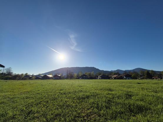 A green meadow, houses and a mountain in the back, the sun is in a blue sky