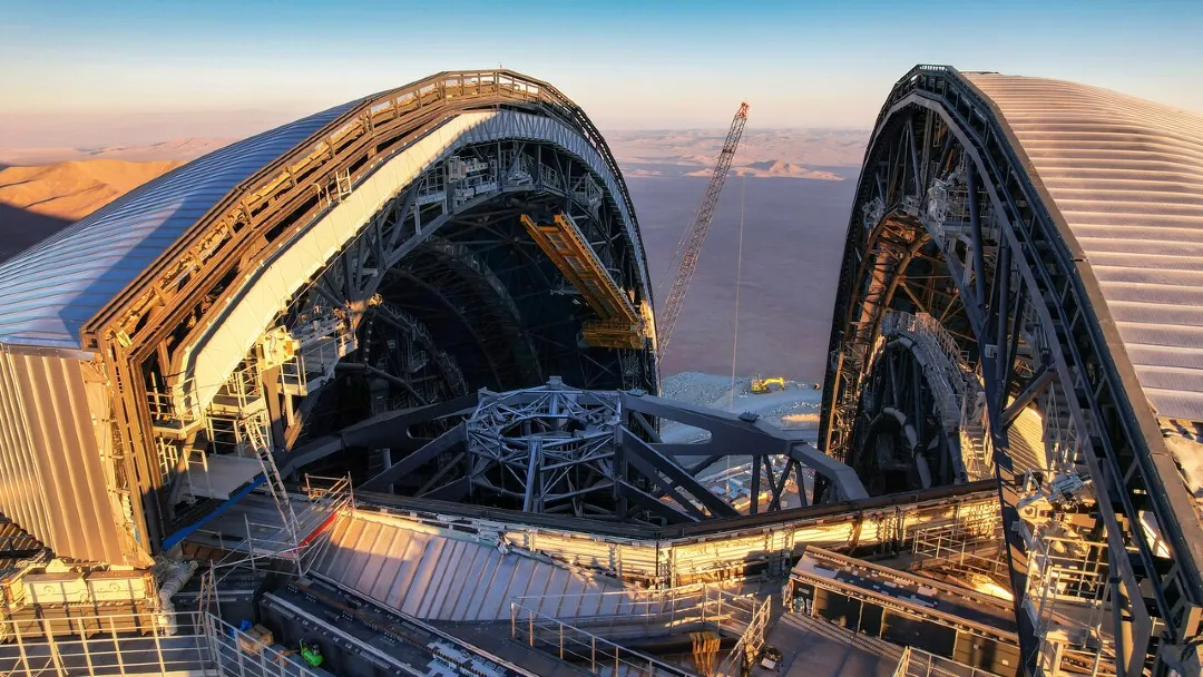 An aerial image showing a huge dome from above, partially split into two halves. Along the edges of each half there’s a complex network of metallic beams and catwalks. The opening reveals part of the metallic frame of a huge telescope inside the dome.