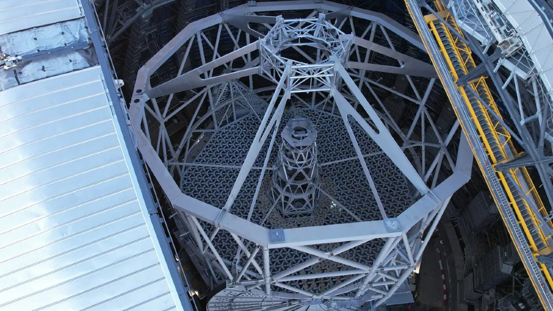 Top-down view of a telescope structure under construction, showing a large circular metal framework with radial support beams and a central tower, surrounded by parts of the observatory building.