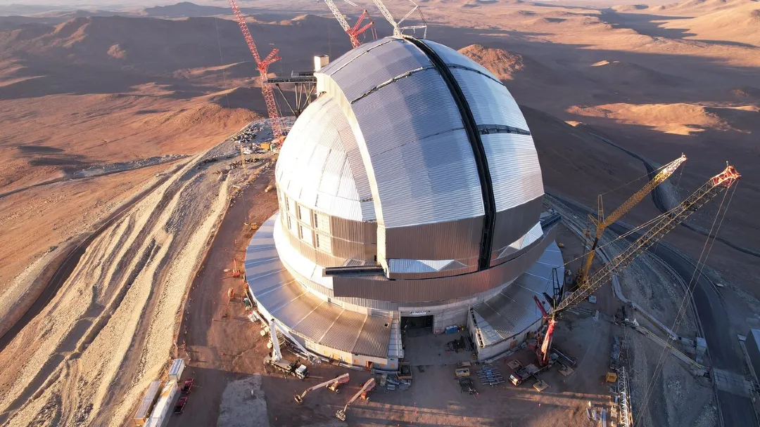 Aerial view of a telescope dome surrounded by cranes and construction equipment, in a desert landscape.