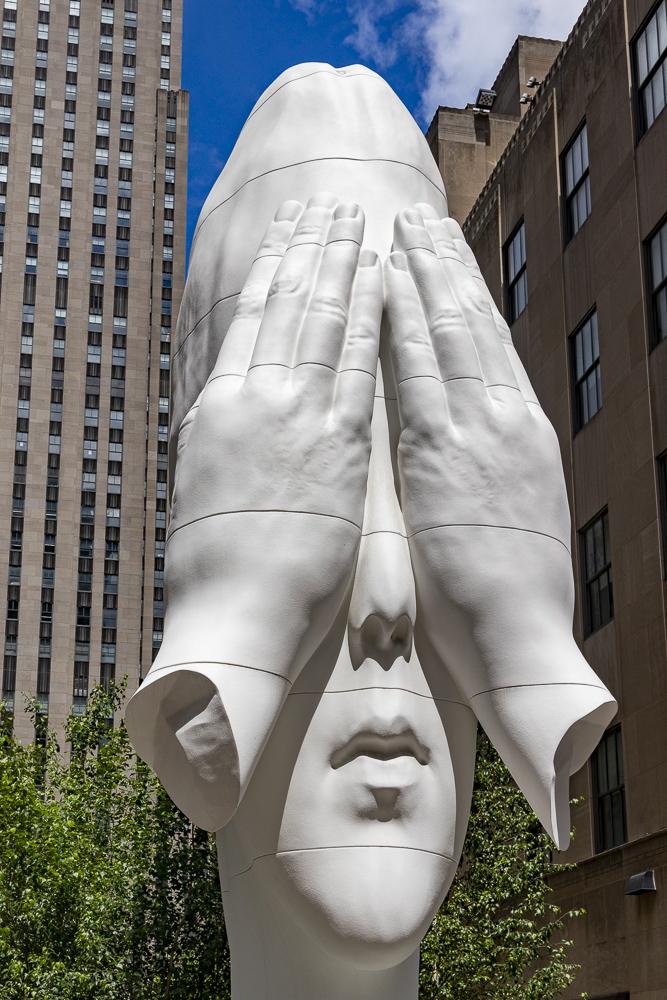 Ein Hochkantfoto zeigt fast formatfüllend eine riesige weiße Skulptur die einen menschlichen Kopf darstellt  mit zwei großen Händen, die das Gesicht bedecken. Das Sonnenlicht verstärkt durch harte Schatten die Struktur. Im Hintergrund sind einige Gebäudeteile des Centers sowie rechts oben in der Eckes etwas blauer leicht bewölkter Himmel zu sehen.