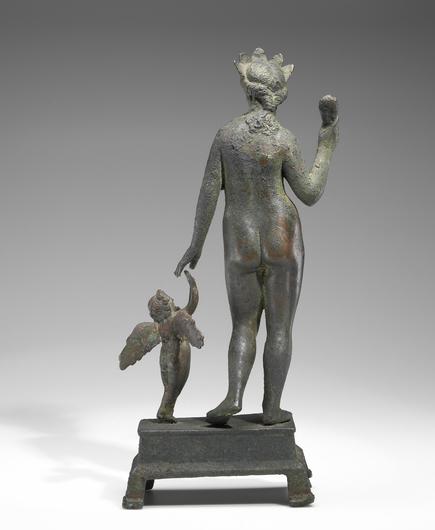 Photo of a bronze sculpture of Aphrodite holding her sandal in a raised right hand, her son Eros standing to her left. Both are seen from behind in this photo. Both are in the nude. Aphrodite wears a crown and her her hair braided. Eros is depicted as a winged child reaching up to his mother.