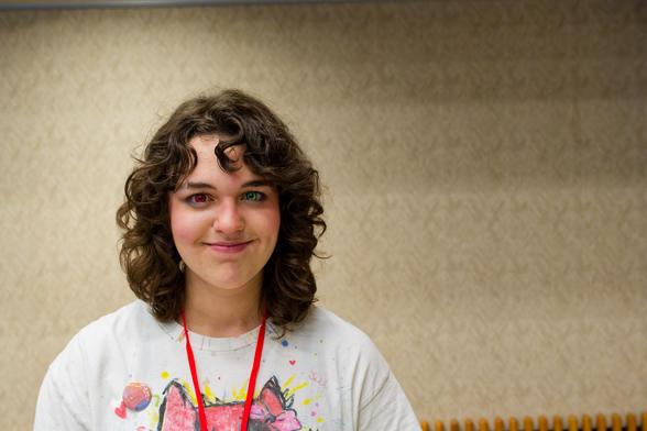 Young woman with auburn drills has the top of a conference lanyard and white shirt with just the ears of a stylized animal showing through with a beige interior hallway behind