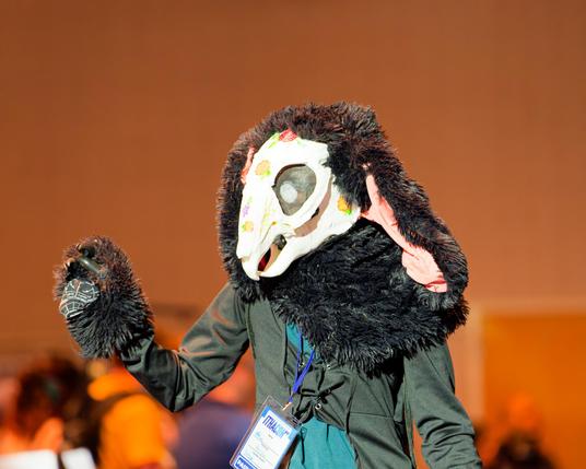 Person with large white skull-like face mask wearing coat and a lot of hair looks and waves to the left