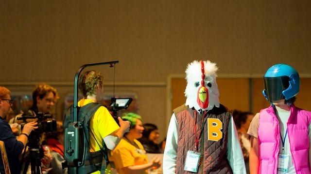 Small and to the left are a person with a chicken head and brown jacket labeled B and a person with a blue helmet with black visor and pink jacket and white shirt abd behind them a man has a fancy harness that holds up him camera with a crowd spectators to his left